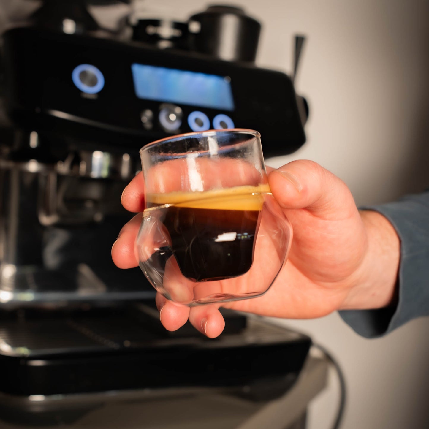 Hand holding a small glass with espresso in front of a coffee machine.