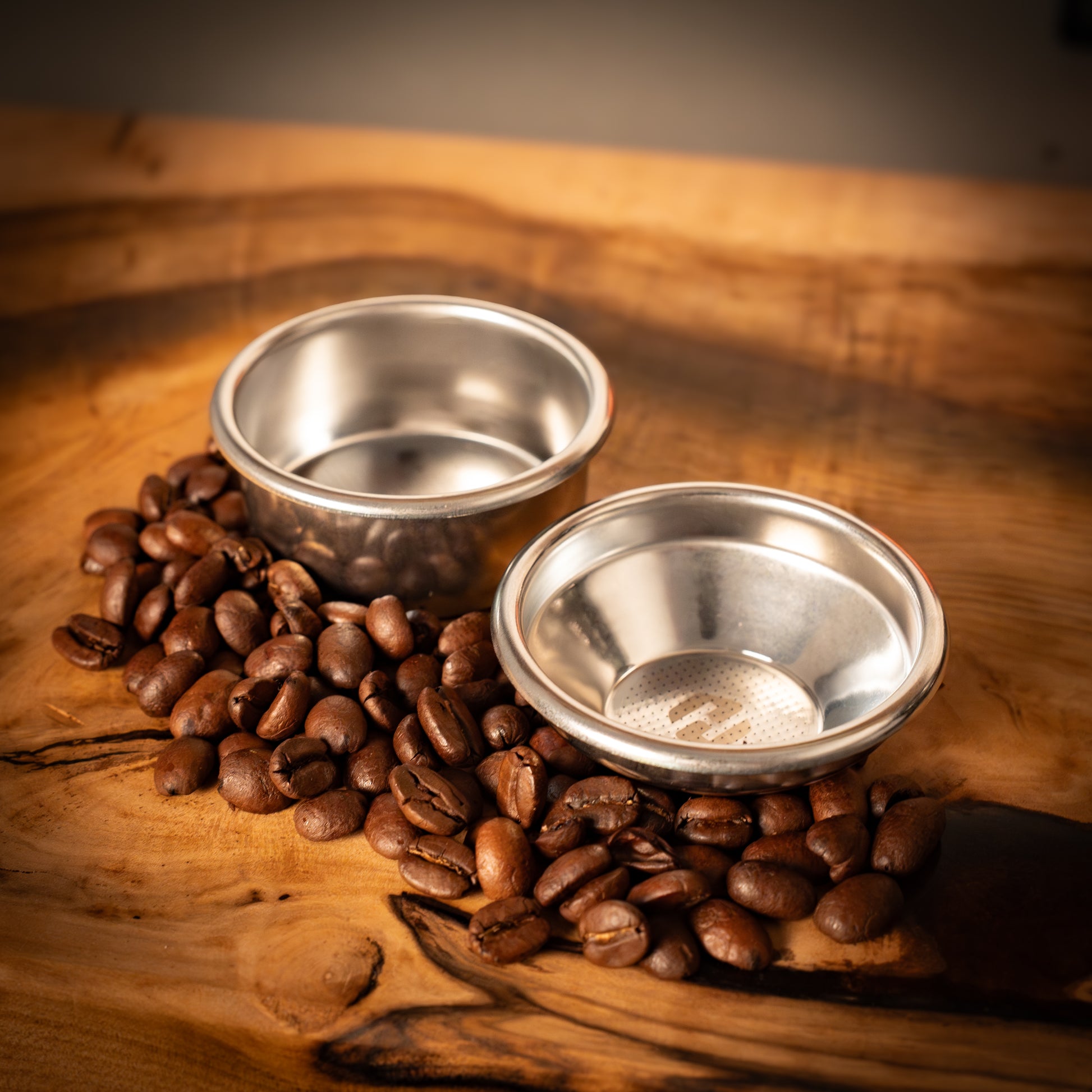 Two portafilter baskets on a wooden surface with coffee beans around them.